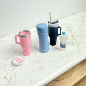 May include: A collection of miniature drinkware items, including a pink tumbler with a handle, a blue thermos, a black travel mug with a white straw, a water bottle, and two clear glasses. A white straw and a small plate with a pink item are also present.