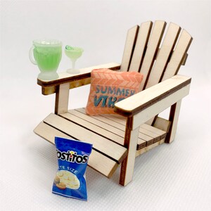 May include: A miniature wooden adirondack chair with a pink pillow that says "Summer Vibes" on it. A small bag of white cheddar cheese flavored tortilla chips and two miniature glasses with green liquid are on the chair's armrests.
