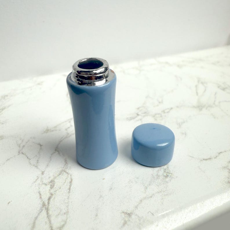 May include: A light blue ceramic bottle with a silver-colored rim and a matching cap. The bottle has a curved shape and is displayed on a white marble surface. The cap is placed next to the bottle.