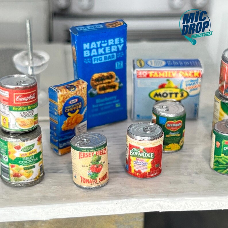 May include: A collection of miniature canned goods, including Campbell's Healthy Request Chicken Noodle Soup, a fruit cocktail can, a can of Jersey Fields Tomato Sauce, a can of Boyardee Mini with cheese and pasta, a can of Mott's Fruit Snacks, and a box of Nature's Bakery Fig Bar.