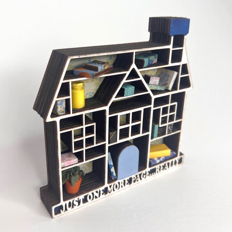 Op de afbeelding: Een houten huisvormig rek met een blauw dak en een witte binnenkant. De planken zijn gevuld met kleine voorwerpen, waaronder boeken, potten en planten. De onderkant van het rek heeft de tekst "Just one more page really".
