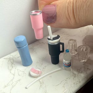 May include: Miniature drinkware set featuring a pink tumbler, a black travel mug with a handle, a blue thermos, a small water bottle, two clear glasses, a straw, and a pink and white lid. The set is arranged on a white and gray marble surface.