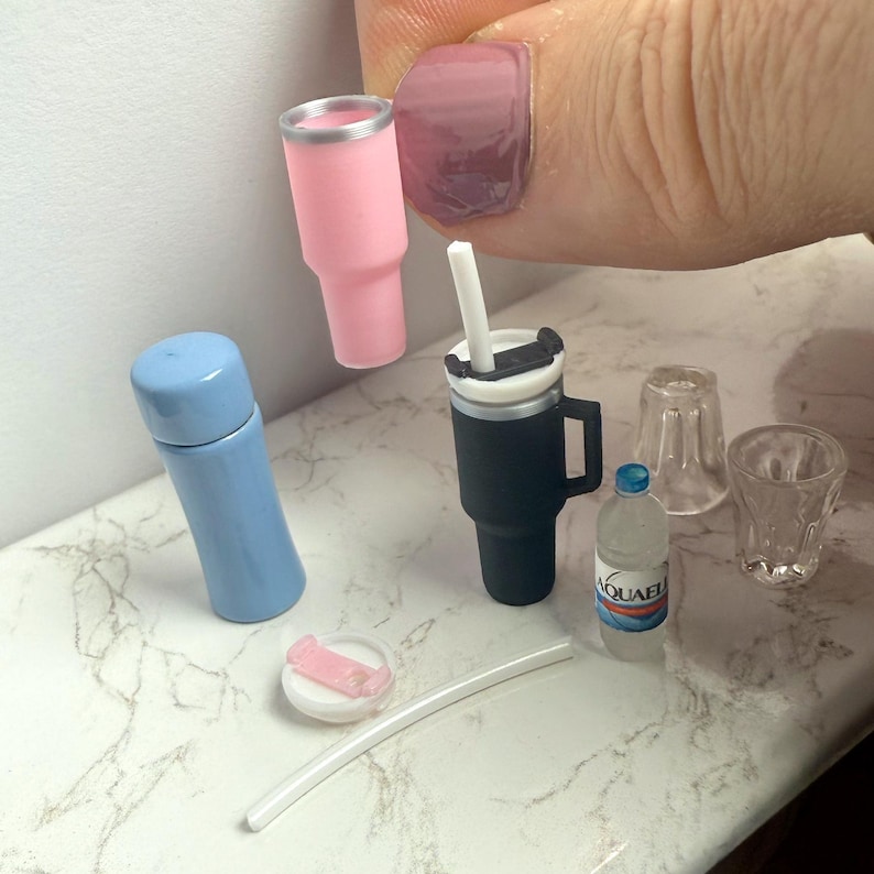 May include: Miniature drinkware set featuring a pink tumbler, a black travel mug with a handle, a blue thermos, a small water bottle, two clear glasses, a straw, and a pink and white lid. The set is arranged on a white and gray marble surface.