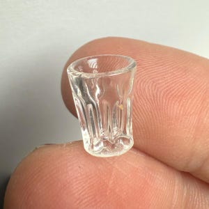 May include: A small, clear glass shot glass with a textured, ridged design. The miniature glass is held between two fingers, showcasing its size and detail. The glass is transparent, allowing light to pass through.