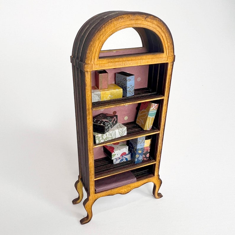 Op de afbeelding: Een houten boekenkast met vier planken, elk gevuld met kleine dozen en boeken. De boekenkast is lichtbruin geverfd en heeft een decoratieve boog aan de bovenkant.