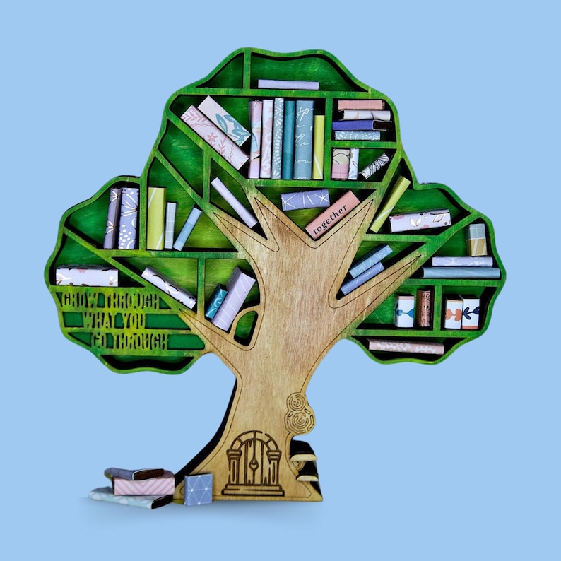 Op de afbeelding: Een houten boom boekenkast met groene bladeren en planken vol kleine boeken. De boomstam heeft een deur aan de basis en de tekst "Grow through what you go through" staat op de stam. De tekst "together" staat op een plank boven de deur.