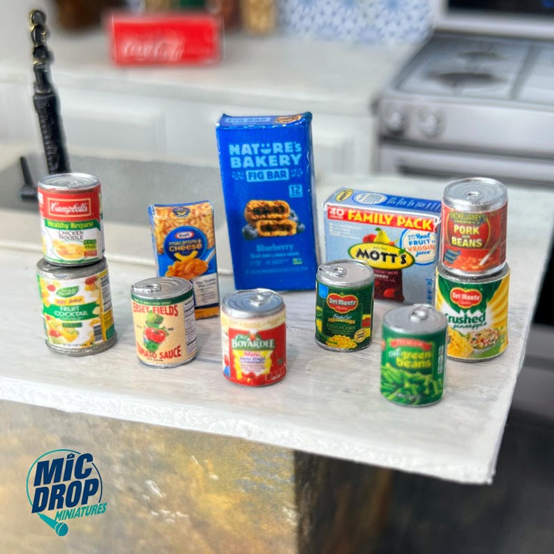 May include: A collection of miniature canned goods, including fruit cocktail, tomato sauce, green beans, pork and beans, crushed fruit cocktail, and a box of macaroni and cheese. There is also a box of blueberry fig bars.