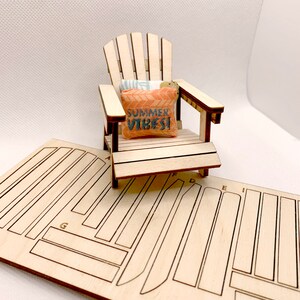 May include: A wooden model of a miniature Adirondack chair with a pillow that says "Summer Vibes!" The chair is sitting on a sheet of wood with cut-out pieces for assembly.