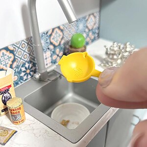 Może przedstawiać: Miniaturowa scena kuchenna z ż&oacute;łtą wyciskarką do cytryn nad zlewem. Pudełko z "Cukrem" i puszka "Sody oczyszczonej" znajdują się na blacie. Zlew jest ze stali nierdzewnej, a backsplash ma niebiesko-biały wz&oacute;r płytek.