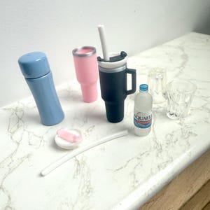 May include: A collection of miniature drinkware items, including a blue thermos, a pink tumbler, a black travel mug with a white straw, a water bottle, two clear glasses, and a white straw with a pink lid.