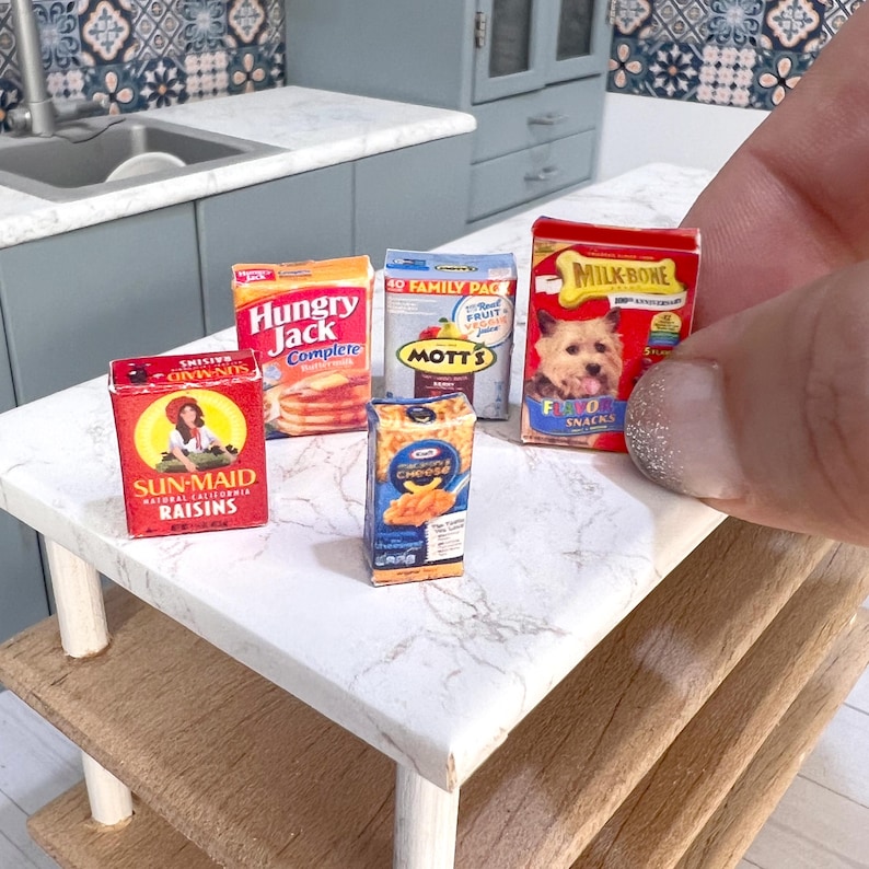 May include: A miniature kitchen countertop with five miniature boxes of cereal, pancake mix, fruit juice, macaroni and cheese, and dog treats. The boxes are on a white marble countertop with a wooden shelf below.