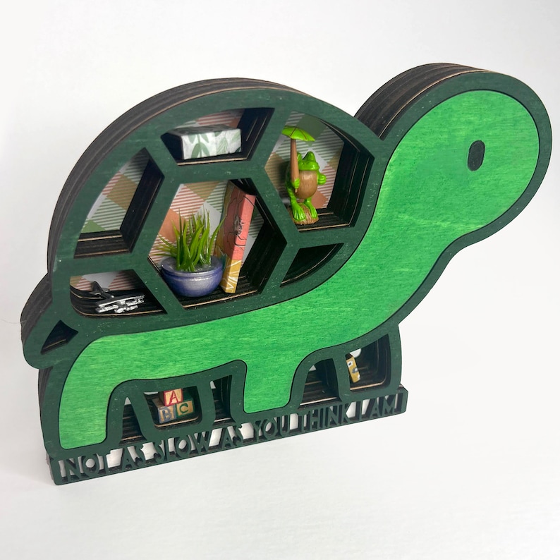 Op de afbeelding: Een groene houten schildpadplank met een zwart interieur en meerdere vakken. De plank heeft een bordje aan de onderkant met de tekst "Niet zo langzaam als je denkt dat ik ben".