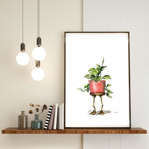 May include: A watercolor painting of a potted plant with green leaves. The pot is pink and has gold legs that look like chicken feet. The painting is framed and hanging on a white wall.