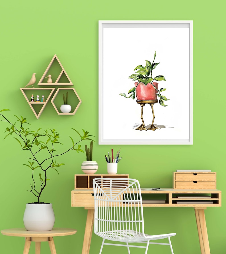 May include: A watercolor illustration of a potted plant with legs. The plant is in a red pot and has green leaves. The plant is standing on two legs with feet. The illustration is framed in a white frame and is hanging on a green wall.