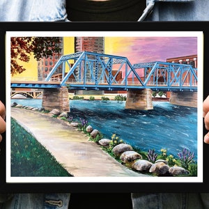 K&ouml;nnte beinhalten: Ein Gem&auml;lde einer blauen Br&uuml;cke &uuml;ber einem Fluss mit einer Stadt im Hintergrund. Der Himmel ist leuchtend rosa und lila. Der Fluss ist blau und es gibt Felsen und gr&uuml;ne Pflanzen am Flussufer.