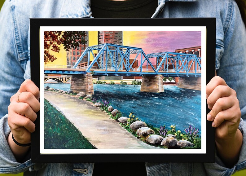 K&ouml;nnte beinhalten: Ein Gem&auml;lde einer blauen Br&uuml;cke &uuml;ber einem Fluss mit einer Stadt im Hintergrund. Der Himmel ist leuchtend rosa und lila. Der Fluss ist blau und es gibt Felsen und gr&uuml;ne Pflanzen am Flussufer.