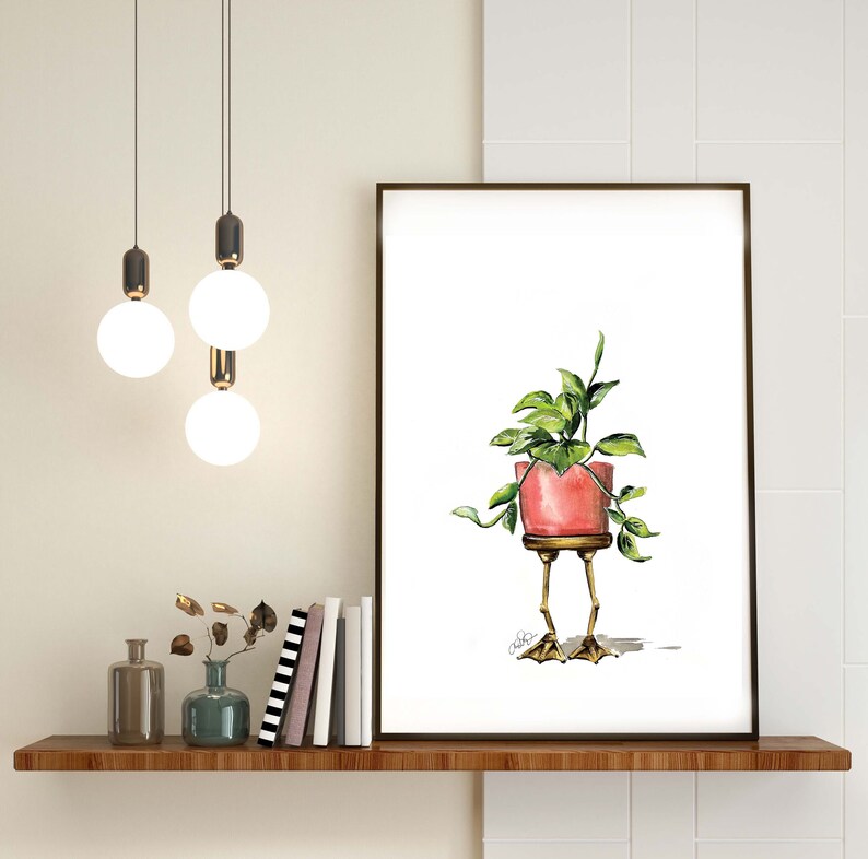 May include: A watercolor painting of a potted plant with green leaves. The pot is pink and has gold legs that look like chicken feet. The painting is framed and hanging on a white wall.