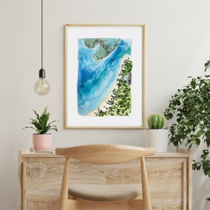 May include: A watercolor painting of a beach scene with blue water, white sand, and green trees. The painting is framed in a light wood frame and is hanging on a white wall.