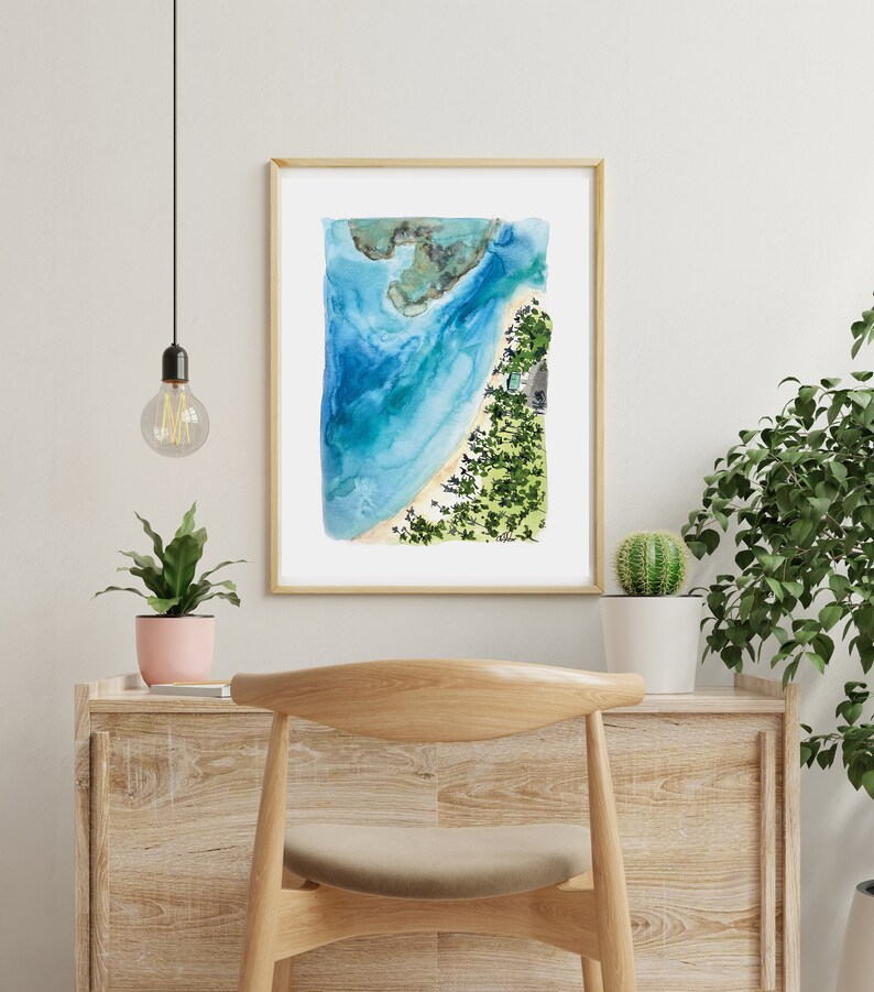 May include: A watercolor painting of a beach scene with blue water, white sand, and green trees. The painting is framed in a light wood frame and is hanging on a white wall.