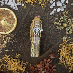 May include: A glass bottle filled with dried herbs, crystals, and a silver bee charm. The bottle is surrounded by dried flowers, orange peel, and other natural elements.