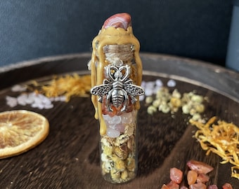 Può includere: Un piccolo flacone di vetro riempito con vari cristalli, fiori secchi ed erbe. Il flacone è sormontato da un sigillo di cera e un ciondolo a forma di ape d'argento. Il flacone è appoggiato su una superficie di legno con fette d'arancia e altri elementi naturali.