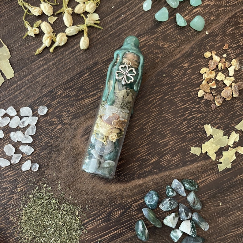 K&ouml;nnte beinhalten: Eine Glasflasche gef&uuml;llt mit gr&uuml;nen und wei&szlig;en Steinen, getrockneten Kr&auml;utern und einem silbernen Gl&uuml;cksbringer in Form eines vierbl&auml;ttrigen Kleeblatts. Die Flasche ist mit gr&uuml;nem Wachs versiegelt und hat einen Korkstopfen.