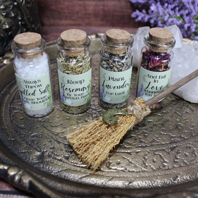 May include: Four small glass jars with cork stoppers containing salt, rosemary, lavender, and dried rose petals. Each jar has a label with text including Always Throw Spilled Salt, Keep Rosemary, Plant Lavender, and And Fall In Love. A small straw broom is included.