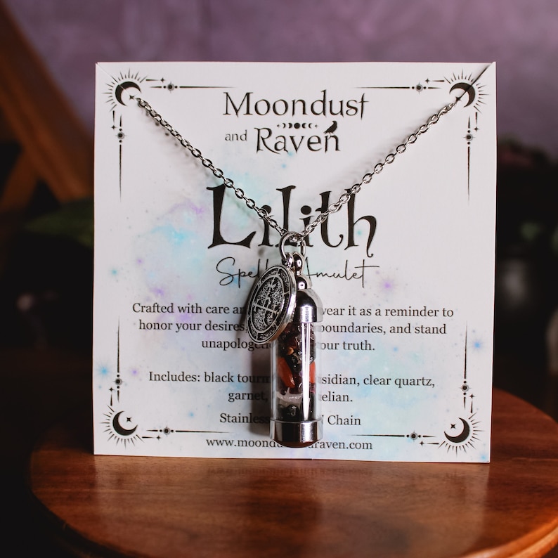 K&ouml;nnte beinhalten: Eine silberne Halskette mit einem Glas-Fl&auml;schchen-Anh&auml;nger, gef&uuml;llt mit schwarzem Turmalin, Granat, Obsidian, klarem Quarz und H&auml;matit. Der Anh&auml;nger ist an einer silbernen Kette befestigt und hat einen silbernen Anh&auml;nger mit einem Pentagramm-Design. Die Halskette wird auf einer wei&szlig;en Karte mit dem Text "Moondust and Raven Lilith Spell Amulet" pr&auml;sentiert.
