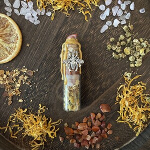 May include: A small glass bottle filled with dried herbs, crystals, and a metal bee charm. The bottle is sealed with a gold wax top and surrounded by dried flowers and other natural elements.