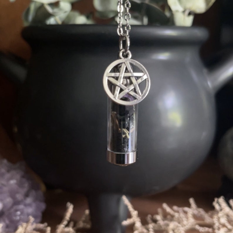 Close-up of a pendant with a silver pentagram charm and a vial containing dried herbs, hanging from a silver chain. Background includes a black cauldron, dried flowers, and an amethyst crystal, creating a mystical, witchcraft-themed ambiance.