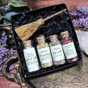 May include: A black gift box containing a small straw broom with a gold moon charm and four glass vials with cork stoppers. The vials contain white salt, dried rosemary, dried lavender, and dried rose petals, each with a printed label.