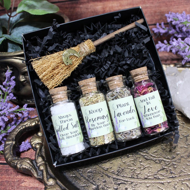 May include: A black gift box containing a small straw broom with a gold moon charm and four glass vials with cork stoppers. The vials contain white salt, dried rosemary, dried lavender, and dried rose petals, each with a printed label.