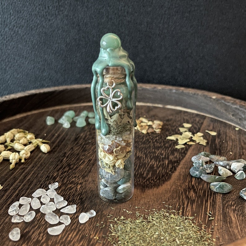 K&ouml;nnte beinhalten: Eine kleine Glasflasche gef&uuml;llt mit verschiedenen gr&uuml;nen und wei&szlig;en Kristallen, Kr&auml;utern und einem Gl&uuml;cksbringer mit vierbl&auml;ttrigem Kleeblatt. Die Flasche ist mit gr&uuml;nem Wachs versiegelt und hat einen silbernen Verschluss.