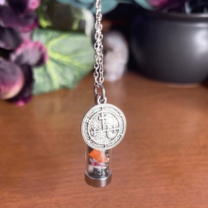 K&ouml;nnte beinhalten: Eine silberne Kettenkette mit einem silbernen Anh&auml;nger mit einem Sigil und einem durchsichtigen Glasfl&auml;schchen, gef&uuml;llt mit kleinen bunten Steinen.