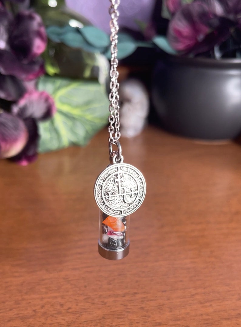 K&ouml;nnte beinhalten: Eine silberne Kettenkette mit einem silbernen Anh&auml;nger mit einem Sigil und einem durchsichtigen Glasfl&auml;schchen, gef&uuml;llt mit kleinen bunten Steinen.