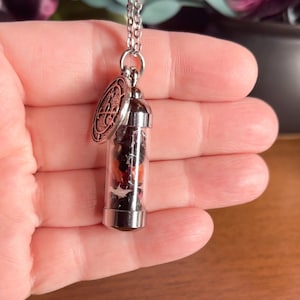 K&ouml;nnte beinhalten: Eine silberne Kettenkette mit einem kleinen Glasfl&auml;schchen-Anh&auml;nger, gef&uuml;llt mit getrockneten Blumen und einem silbernen Anh&auml;nger mit einem Design.