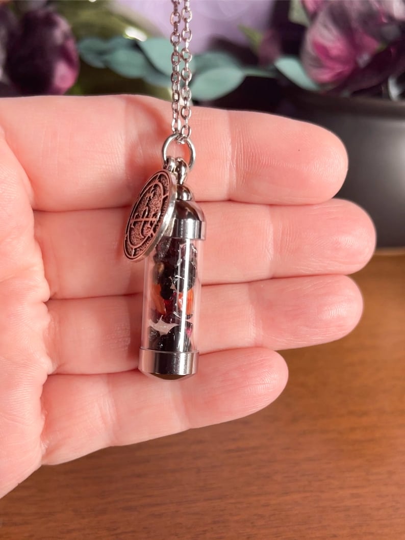 K&ouml;nnte beinhalten: Eine silberne Kettenkette mit einem kleinen Glasfl&auml;schchen-Anh&auml;nger, gef&uuml;llt mit getrockneten Blumen und einem silbernen Anh&auml;nger mit einem Design.