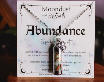 May include: A silver necklace with a glass vial pendant filled with crystals, moss agate, cinnamon, aventurine, pyrite, and gold flakes. The pendant is attached to a silver chain and has a four-leaf clover charm. The card reads "Moondust and Raven Abundance Spell Jarlet".