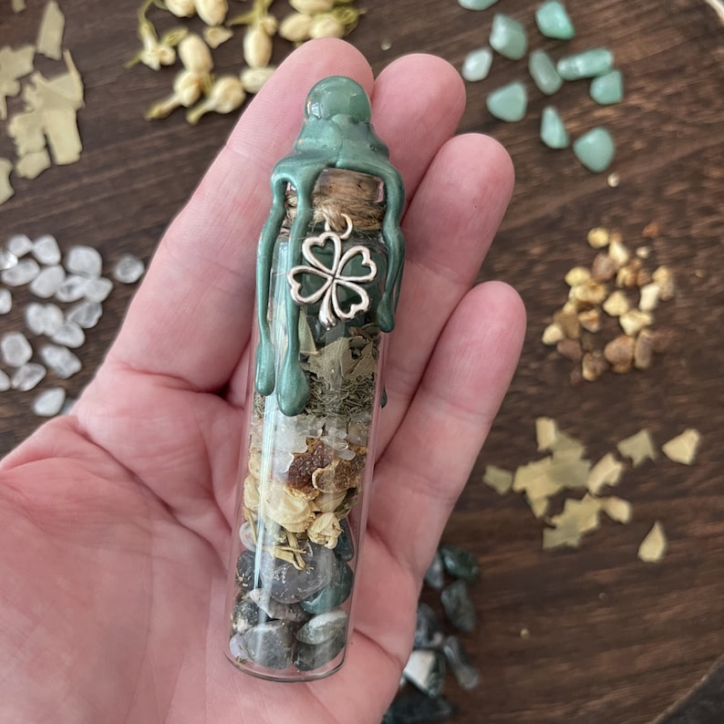 K&ouml;nnte beinhalten: Eine kleine Glasflasche mit einem gr&uuml;nen Wachssiegel und einem Gl&uuml;ckskleeblatt-Anh&auml;nger. Die Flasche ist gef&uuml;llt mit verschiedenen Kr&auml;utern, Kristallen und anderen nat&uuml;rlichen Elementen.