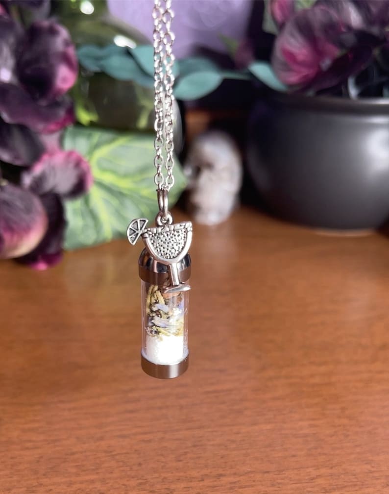 Puede incluir: Un collar de cadena plateada con un peque&ntilde;o colgante de frasco de vidrio. El frasco est&aacute; lleno de hierbas y flores secas y rematado con un dije plateado de un vaso de margarita con una rodaja de lima.