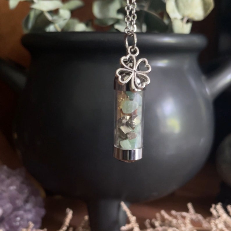 Close-up of a pendant featuring a four-leaf clover charm and a vial containing small green and gold stones, hanging from a silver chain. The background includes a black cauldron, dried flowers, and an amethyst crystal, creating a mystical ambiance.