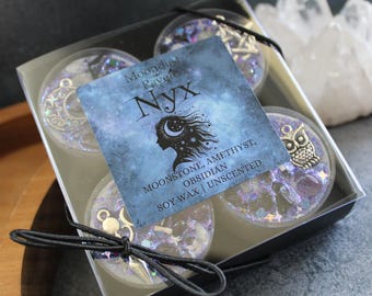 May include: A set of four soy wax candles in a clear plastic box. The candles are decorated with glitter, charms, and crystals. The box has a black background and a label that reads "Moondust and Raven Nyx Moonstone, Amethyst, Obsidian Soy Wax | Unscented".