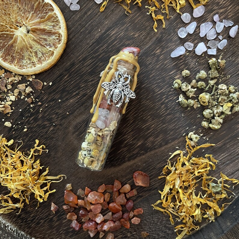 May include: A small glass bottle filled with dried flowers, crystals, and a silver bee charm. The bottle is sealed with a cork and has a golden wax seal. The bottle is sitting on a wooden surface with other natural elements.