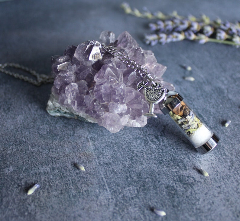 Puede incluir: Un collar de plata con un dije que parece un vaso de c&oacute;ctel lleno de hierbas y flores secas. El collar est&aacute; descansando sobre un grupo de cristales de amatista.