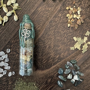K&ouml;nnte beinhalten: Eine gr&uuml;ne Glasflasche mit einem Wachssiegel und einem silbernen Gl&uuml;ckskleeblatt-Anh&auml;nger. Die Flasche ist gef&uuml;llt mit verschiedenen Kristallen, Kr&auml;utern und anderen Pflanzen.