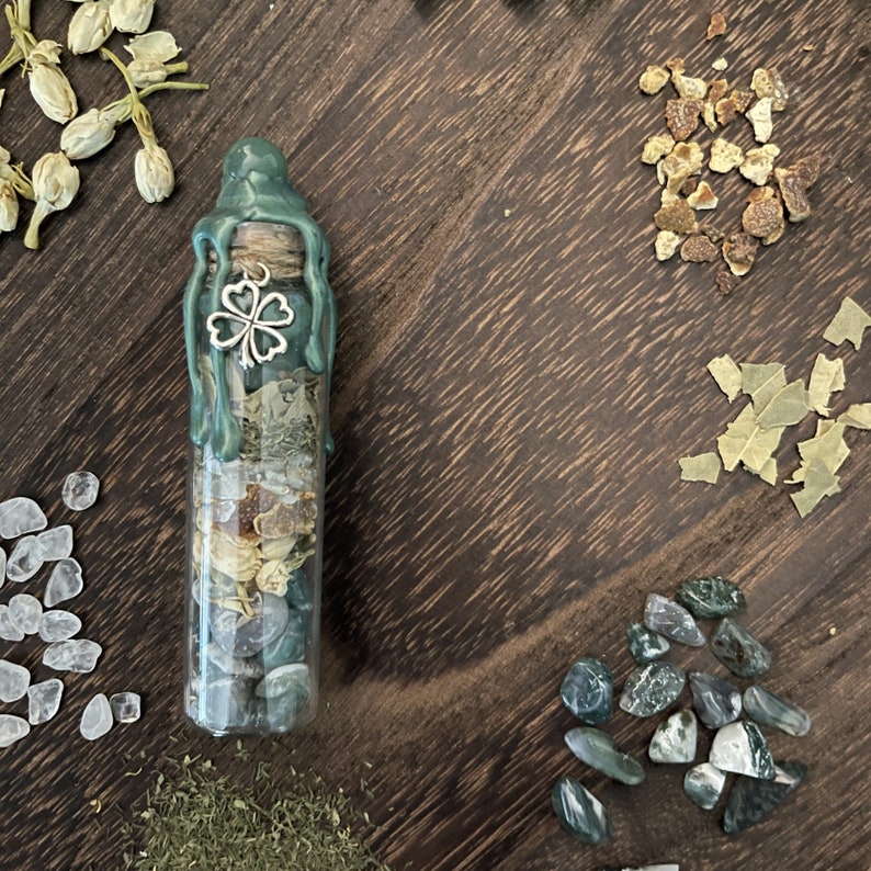 K&ouml;nnte beinhalten: Eine gr&uuml;ne Glasflasche mit einem Wachssiegel und einem silbernen Gl&uuml;ckskleeblatt-Anh&auml;nger. Die Flasche ist gef&uuml;llt mit verschiedenen Kristallen, Kr&auml;utern und anderen Pflanzen.