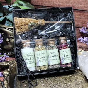 May include: A black gift box containing a small straw broom and four glass vials with cork stoppers. The vials contain white salt, dried rosemary, dried lavender, and pink flower petals, each with labels featuring text and moon icons.