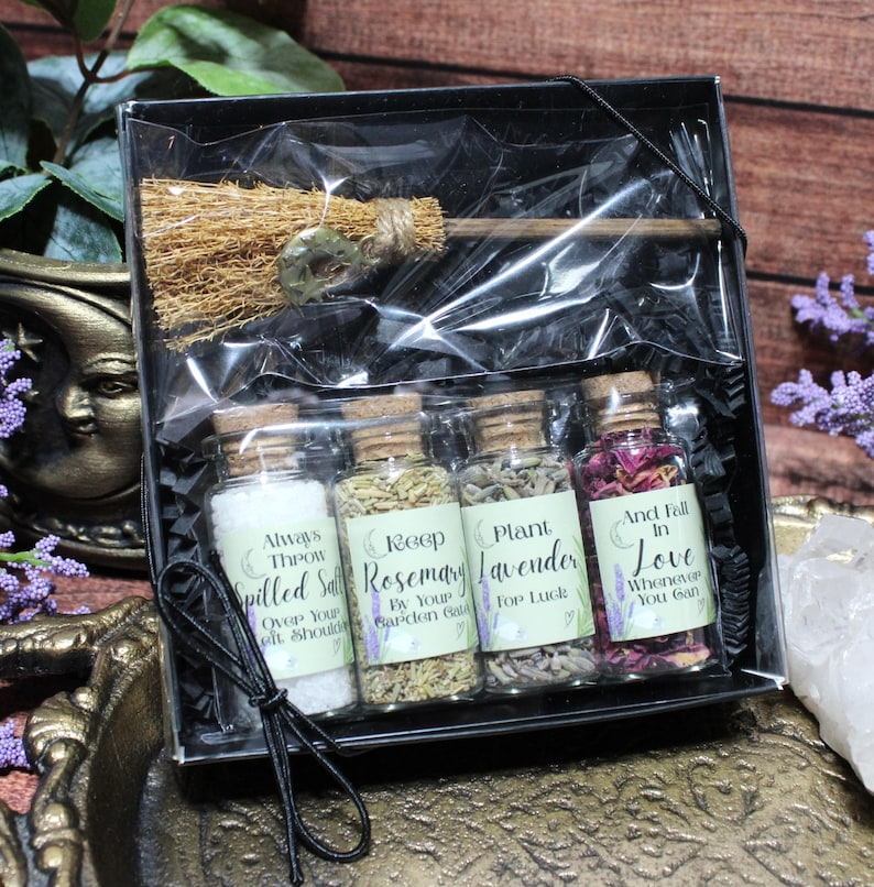 May include: A black gift box containing a small straw broom and four glass vials with cork stoppers. The vials contain white salt, dried rosemary, dried lavender, and pink flower petals, each with labels featuring text and moon icons.