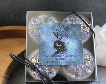 May include: A set of four soy wax candles in a clear plastic box. The candles are unscented and have a celestial theme with embedded glitter and charms. The label reads "Moondust and Raven Nyx", "Moonstone, Amethyst, Obsidian", "Soy Wax | Unscented".