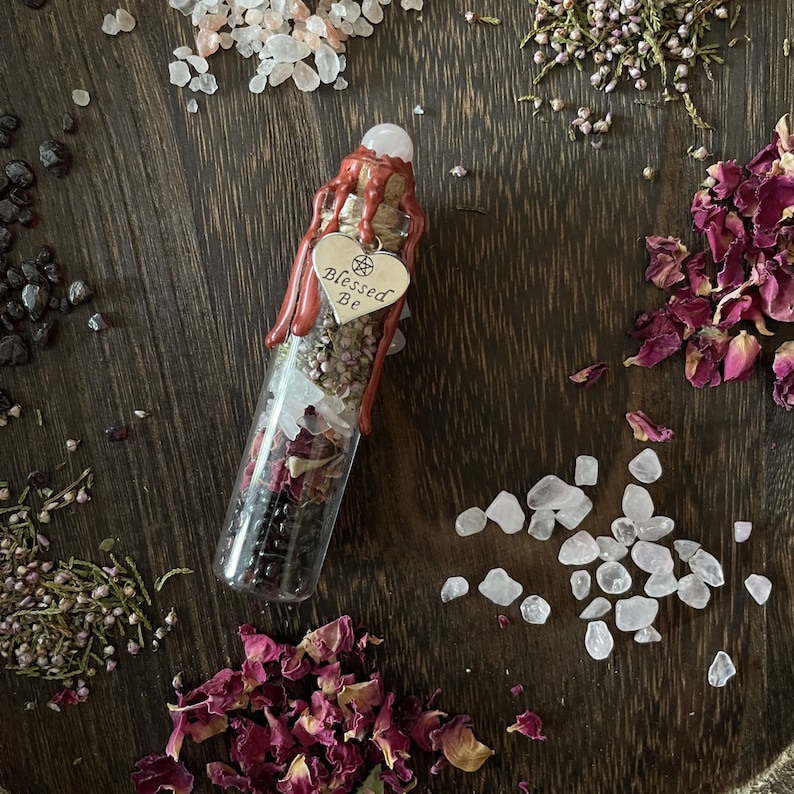 May include: A glass bottle filled with dried herbs, rose petals, and crystals, sealed with red wax and a pentagram charm. The bottle has a silver heart charm with the words "Blessed Be" engraved on it.